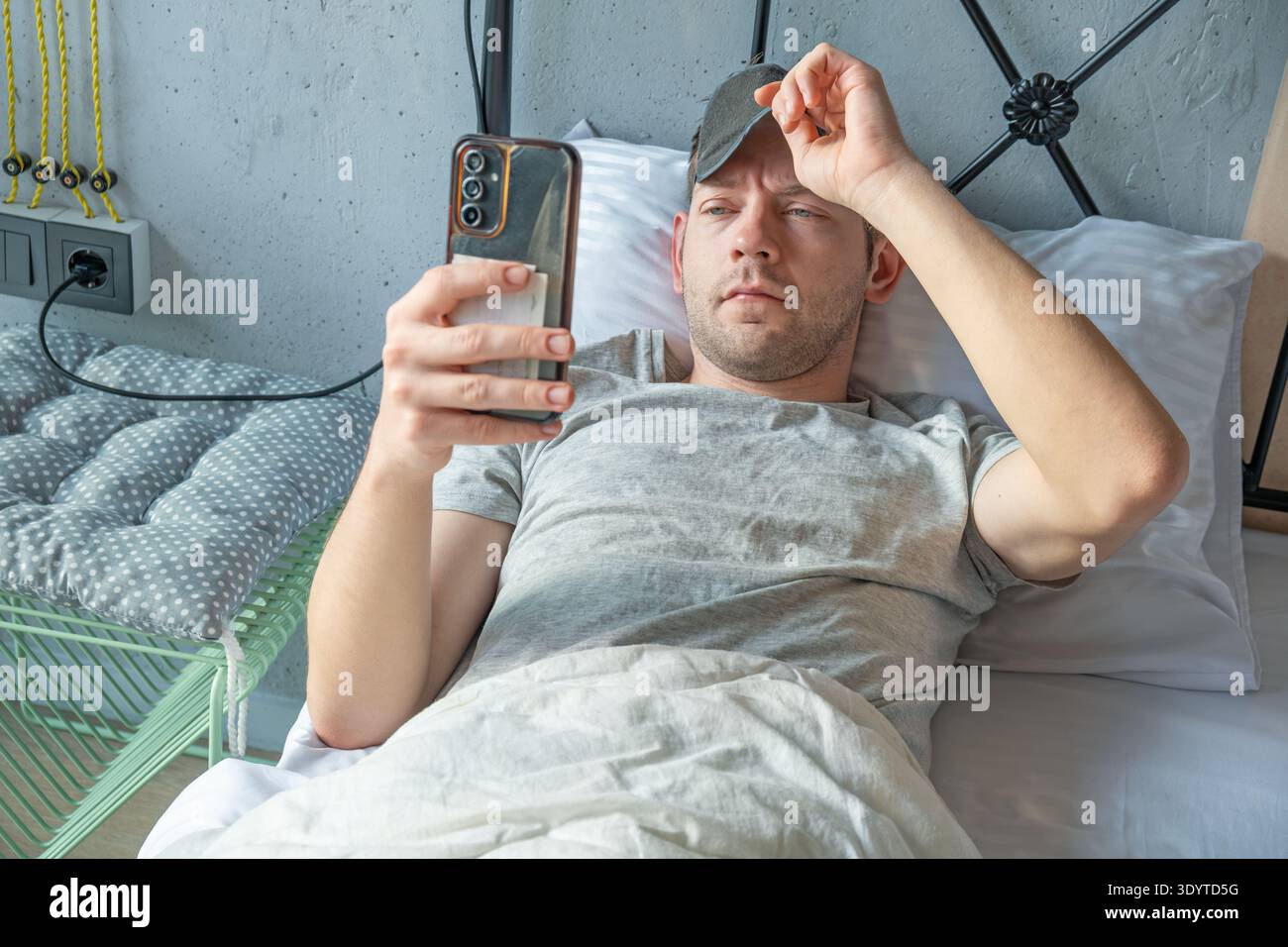Homme allongé dans le lit, enlevant son masque de sommeil pour vérifier les nouvelles sur son smartphone avec des expressions fatigue et concentration comme il vérifie les messages après le réveil Banque D'Images