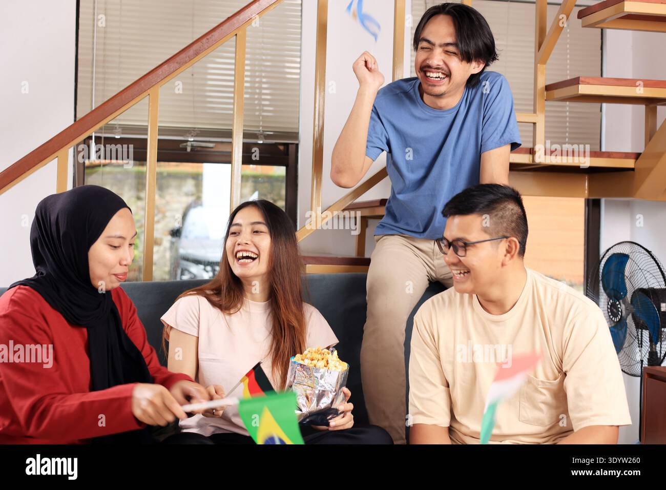 Groupe de jeunes amis asiatiques acclamant tout en regardant un match de sport ensemble, portant différents drapeaux nationaux et montrant l'excitation comme les fans pendant Banque D'Images