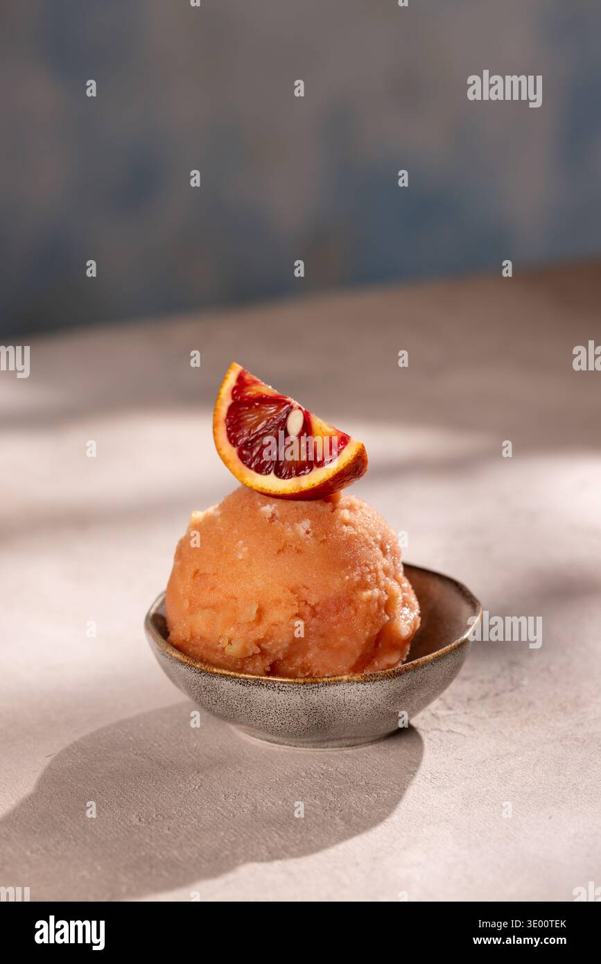 Gros plan d'une boule de sorbet à l'orange sanguine dans un petit bol en céramique, garni d'une tranche d'orange ; entouré de quartiers et d'oranges mûres entières dans un W. Banque D'Images