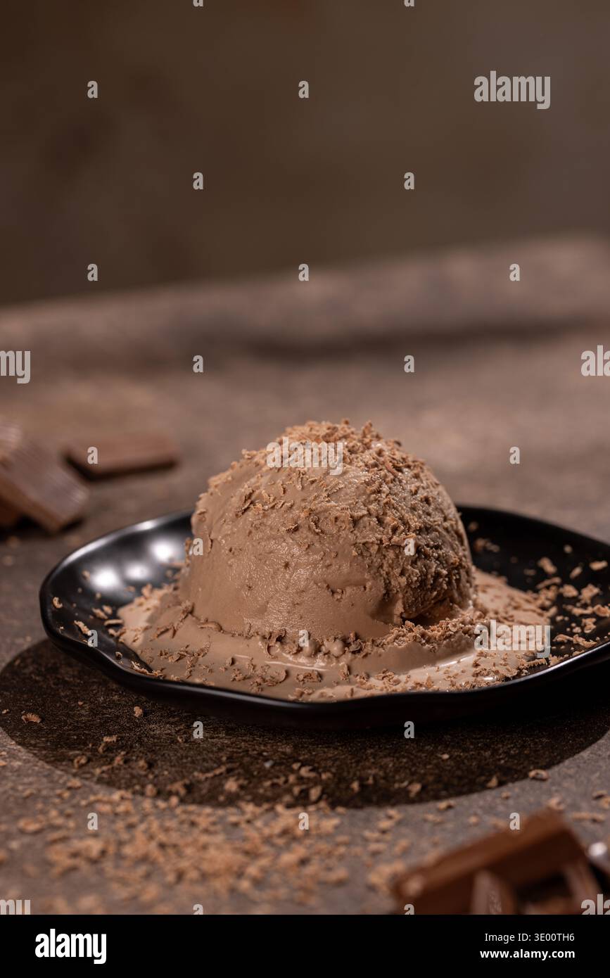 Gros plan d'une grande boule de crème glacée au chocolat crémeuse, saupoudrée de copeaux de chocolat fins, sur une assiette noire mate. Le studio stylé tourné à la guerre Banque D'Images