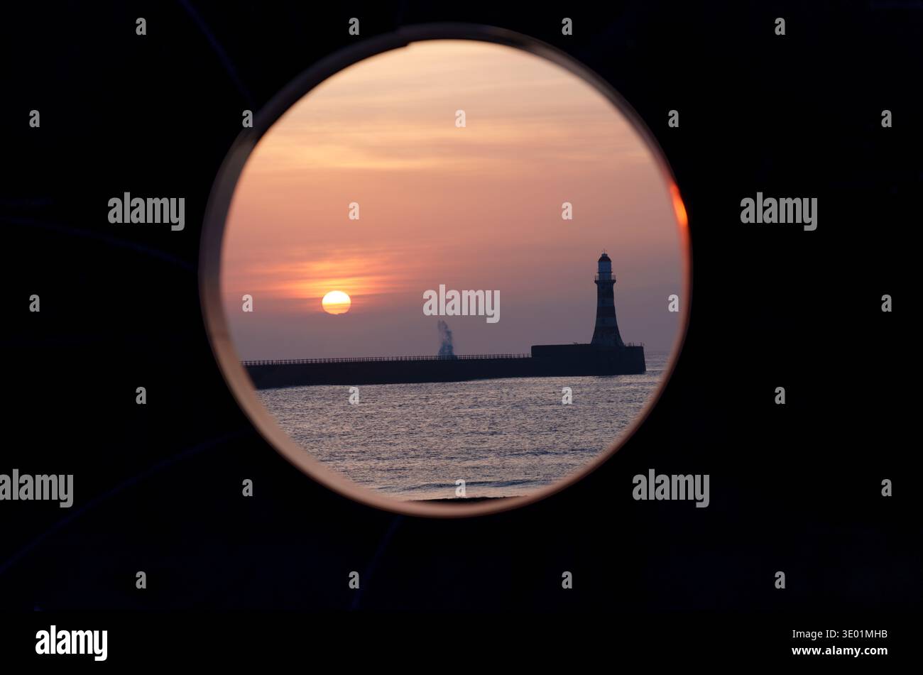 Une composition créative du phare de Roker et du soleil levant, vue à travers l'ouverture circulaire du monume de la route cyclable C2C (Coast to Coast) Banque D'Images