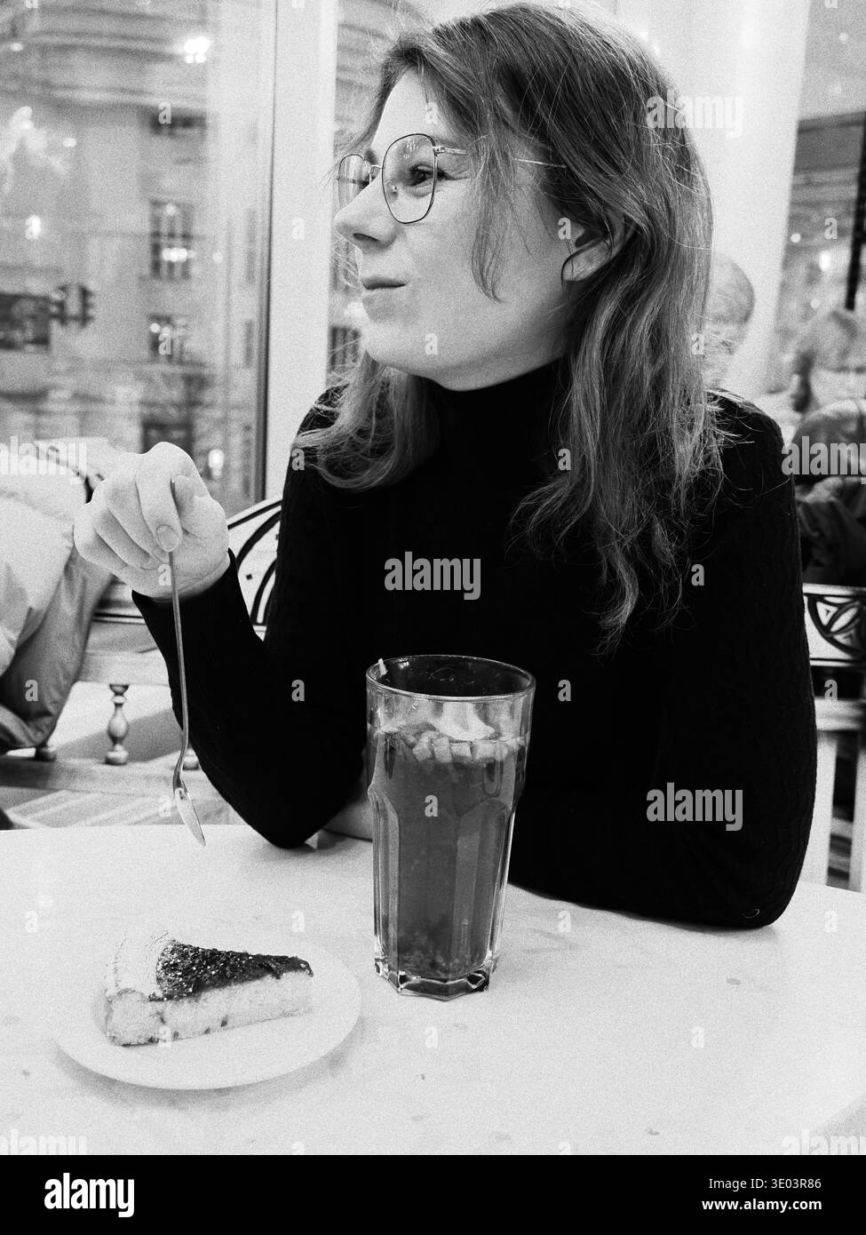 Portrait en noir et blanc d'une femme en lunettes assise dans un café à Tallinn, Estonie, avec une tranche de gâteau et une boisson chaude sur la table. Banque D'Images