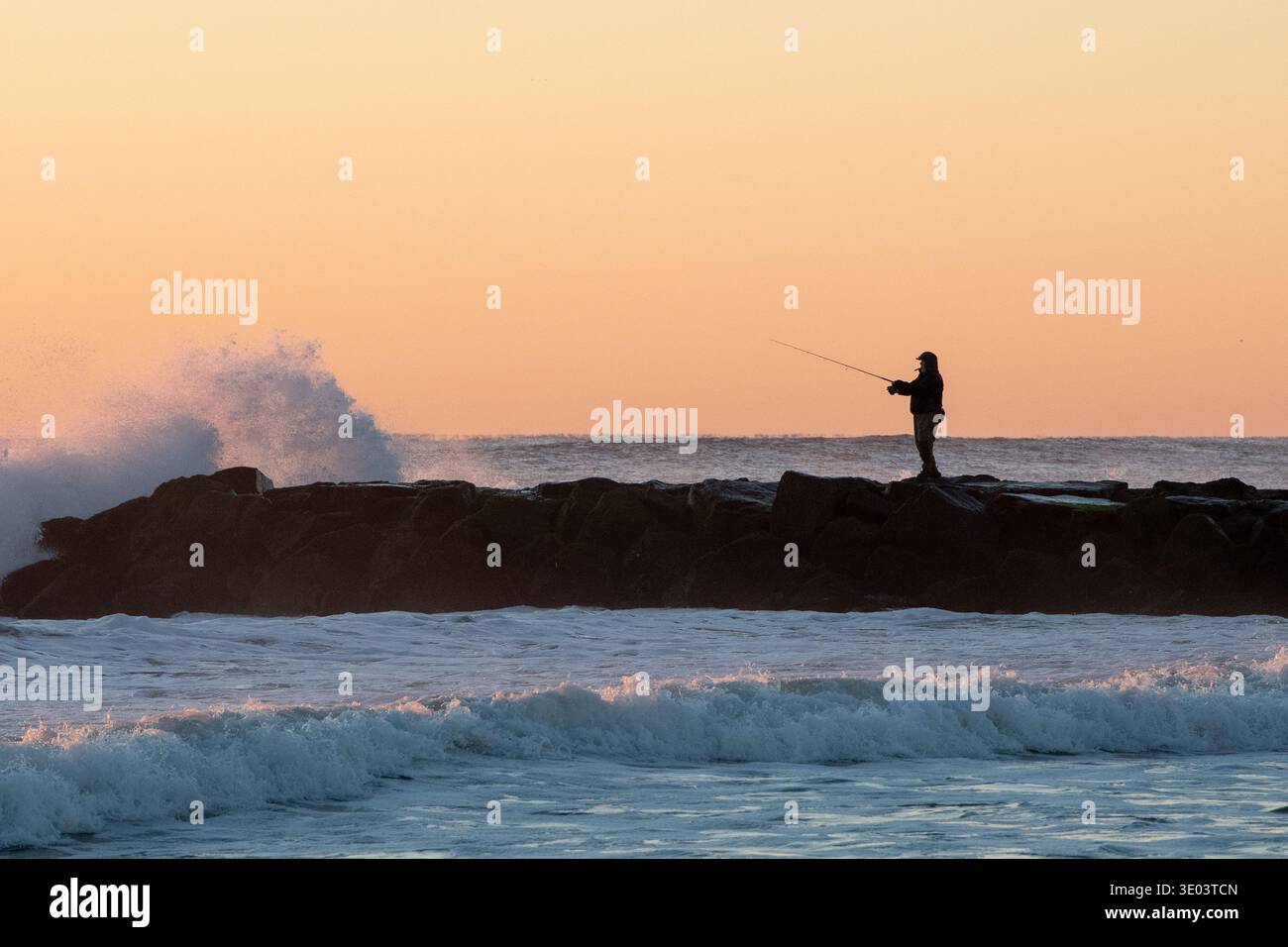 Silhouette d'un pêcheur debout sur une jetée rocheuse alors que les vagues s'écrasent dramatiquement contre elle au lever du soleil, projetant une lueur orange chaude à travers le ciel Banque D'Images
