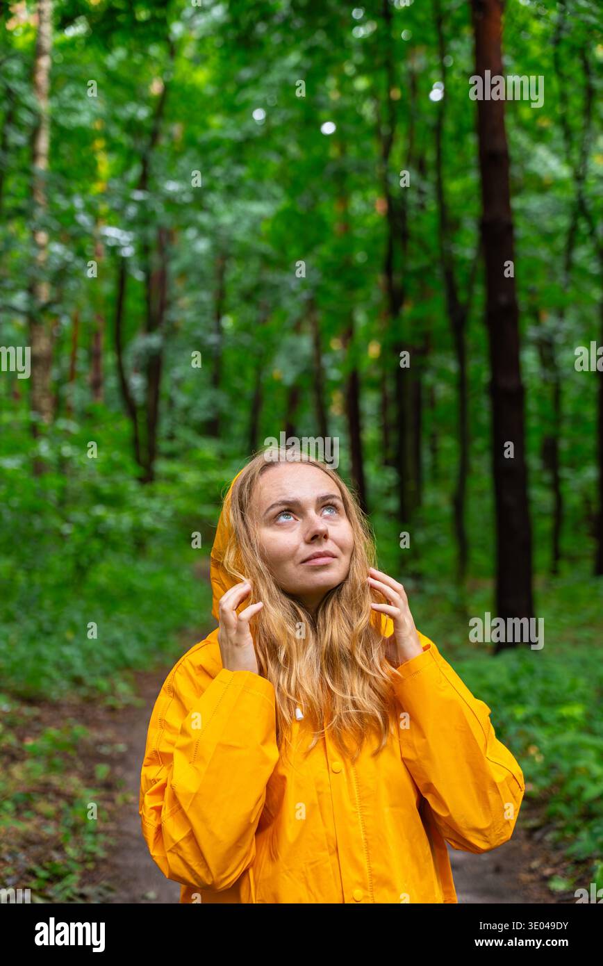 Une femme portant un imperméable jaune vif se tient dans une forêt luxuriante et verte. Elle regarde vers le haut, peut-être à l'écoute des oiseaux, embrassant l'ambiance tranquille i. Banque D'Images