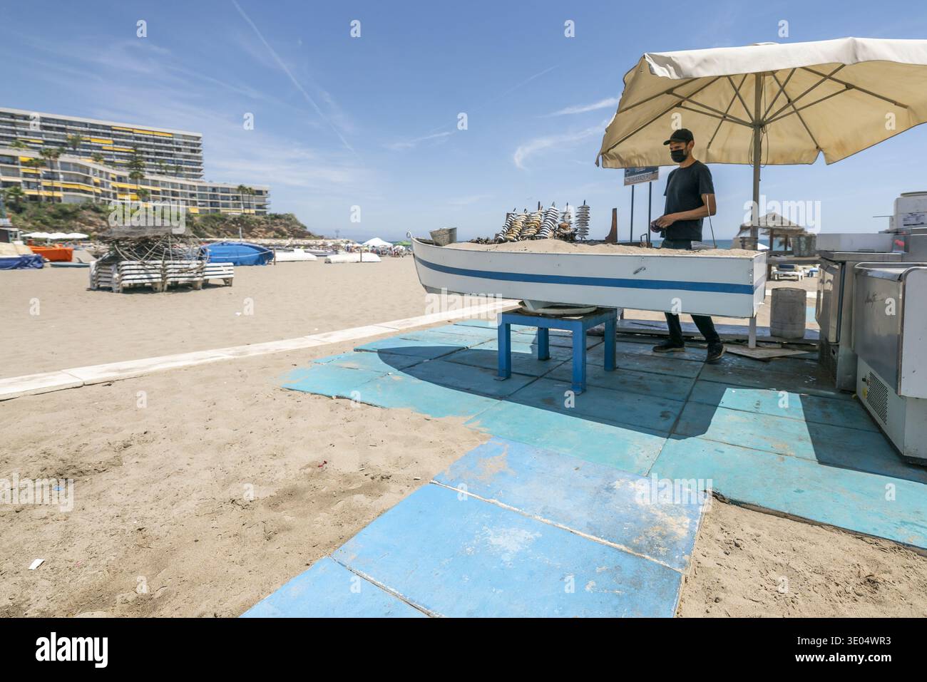 Un vendeur prépare des sardines sur des brochettes dans un stand de nourriture en bord de mer sous un parasol, avec une plage de sable et des hôtels en arrière-plan. Banque D'Images