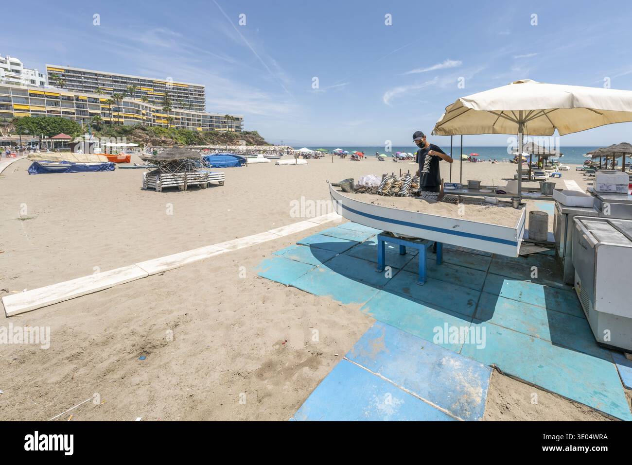 Vendeur de rue griller des sardines sur des brochettes en bois dans un stand de nourriture en bord de mer sous un parasol, avec des bâtiments de plage et de villégiature en arrière-plan. Banque D'Images