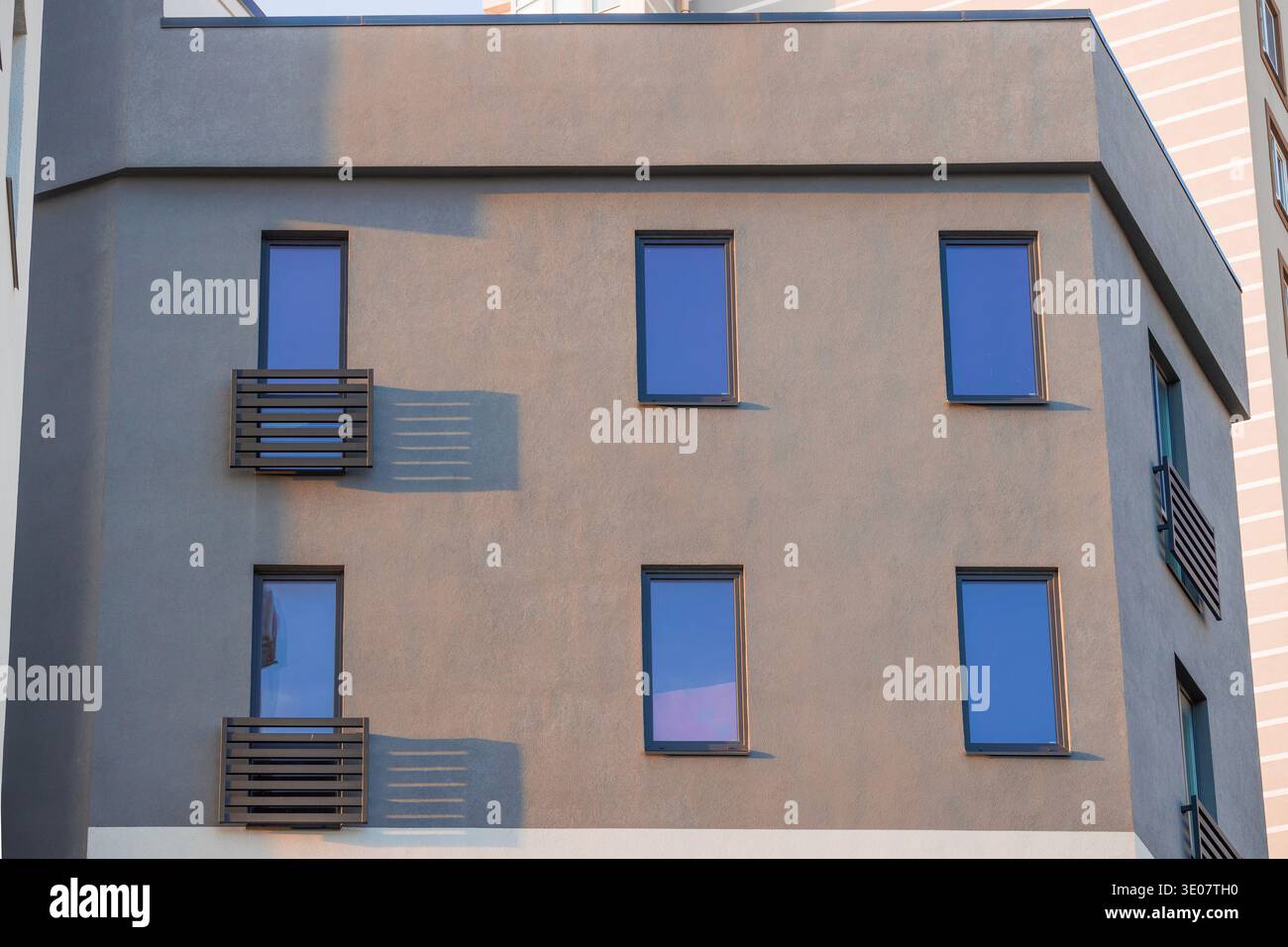 L'extérieur d'un immeuble résidentiel contemporain présente une façade grise texturée avec de multiples fenêtres teintées de bleu et des balcons minimalistes baignés Banque D'Images