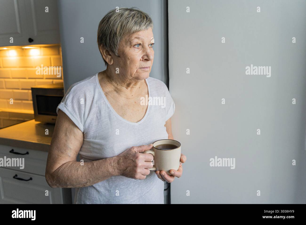 Femme senior regardant par la fenêtre lumineuse, tenant une tasse à café, pensant à la vie ou à l'avenir dans un moment calme Banque D'Images