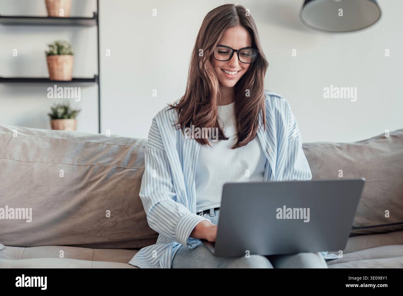 Une jeune femme est assise sur le canapé portant des lunettes, concentrée sur son smartphone. Elle travaille, navigue et fait des achats en ligne à la maison, reflétant la concentration, pro Banque D'Images