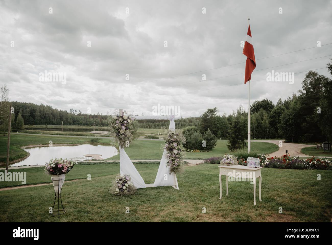 Cadre pittoresque de cérémonie de mariage en plein air avec une arche florale décorée, une table élégante avec boîte à cartes et des arrangements de bouquet sur une pelouse verte près d'un lak Banque D'Images