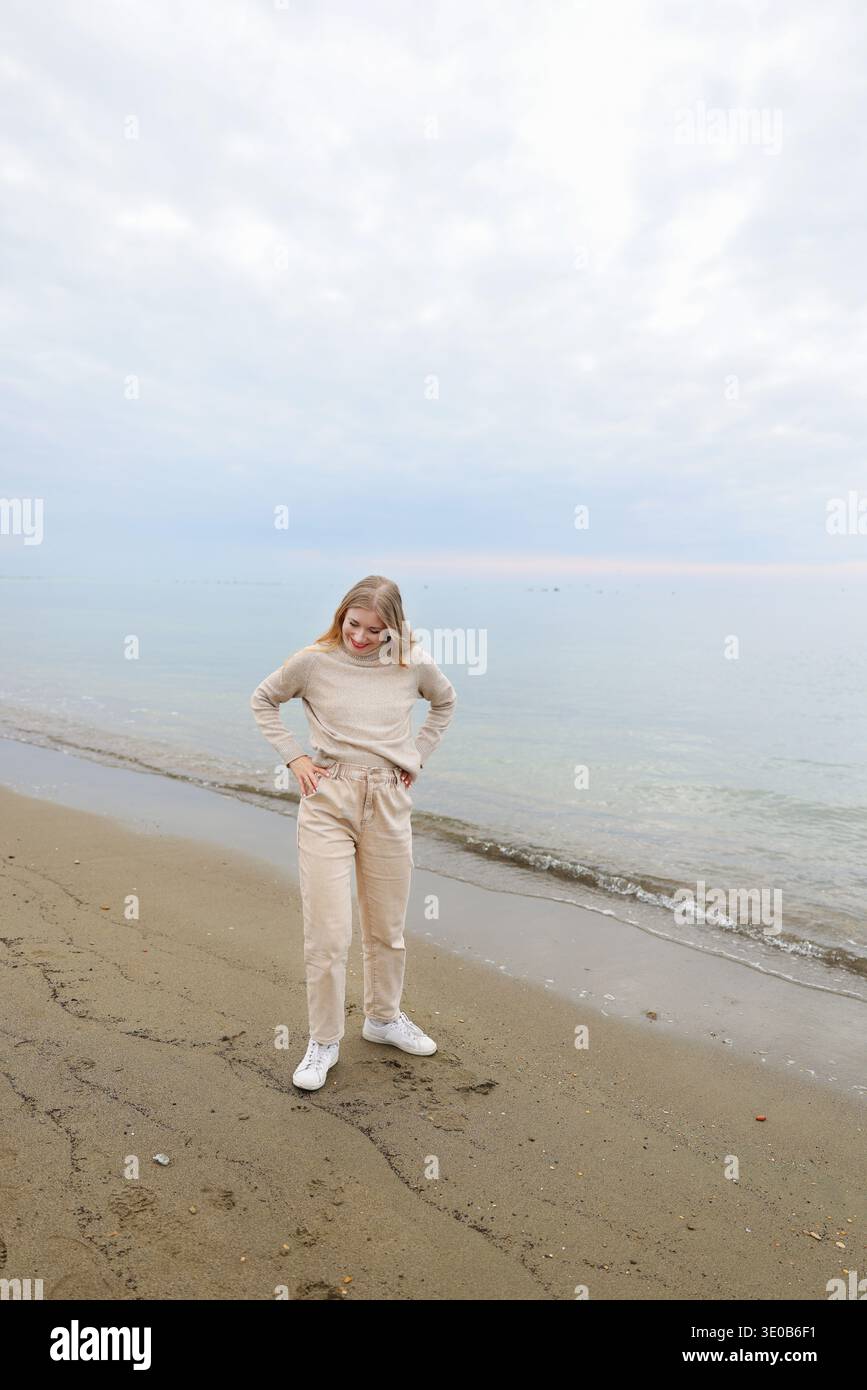 Jeune femme debout sur une plage de sable avec les bras levés vers la mer calme. Liberté et moment paisible de style de vie côtier. Banque D'Images