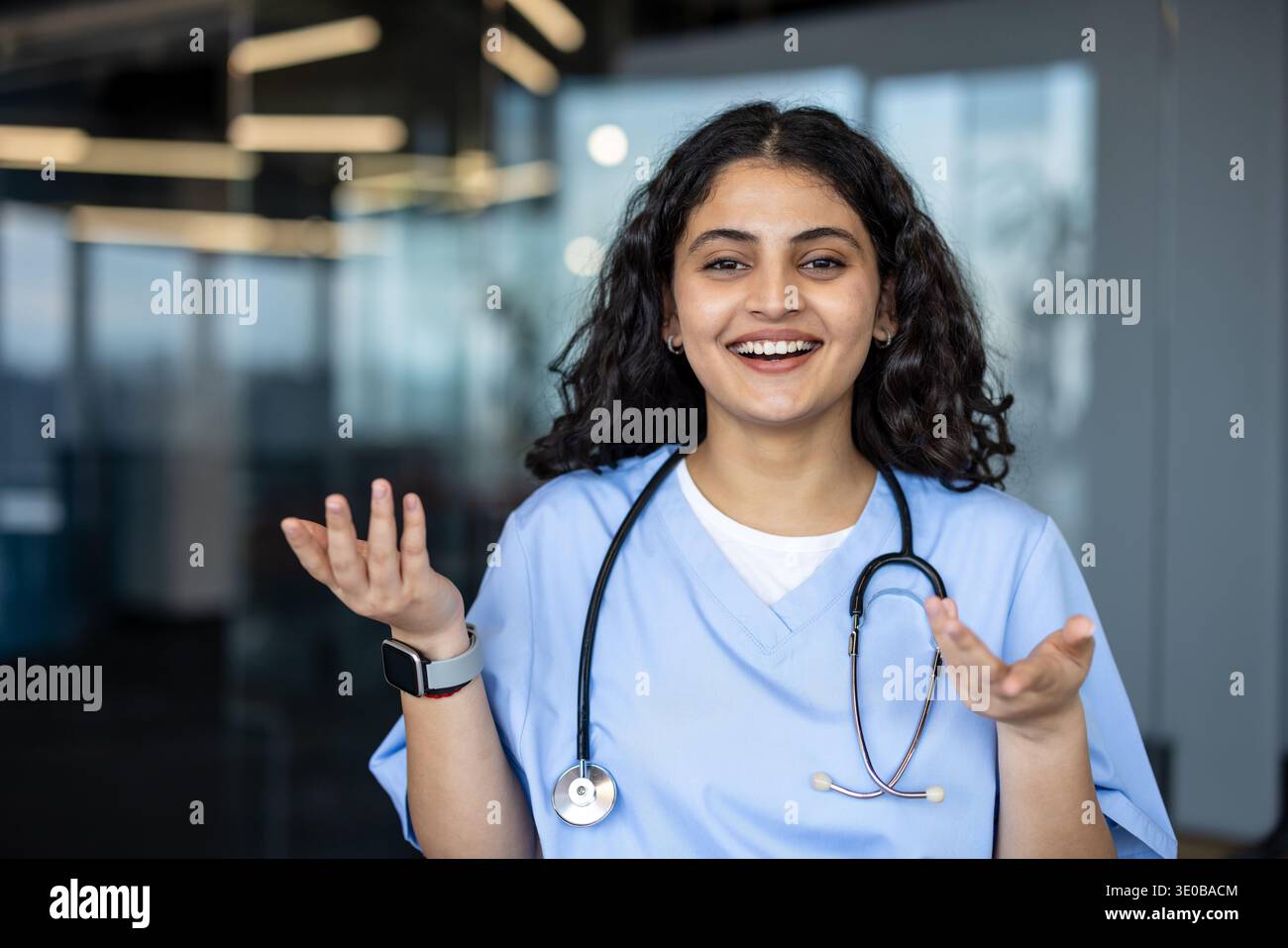 Jeune professionnelle médicale indienne dans les gommages bleus et stéthoscope souriant, parlant et faisant des gestes avec les mains, symbolisant la communication, les soins et le professionnalisme en matière de soins de santé Banque D'Images