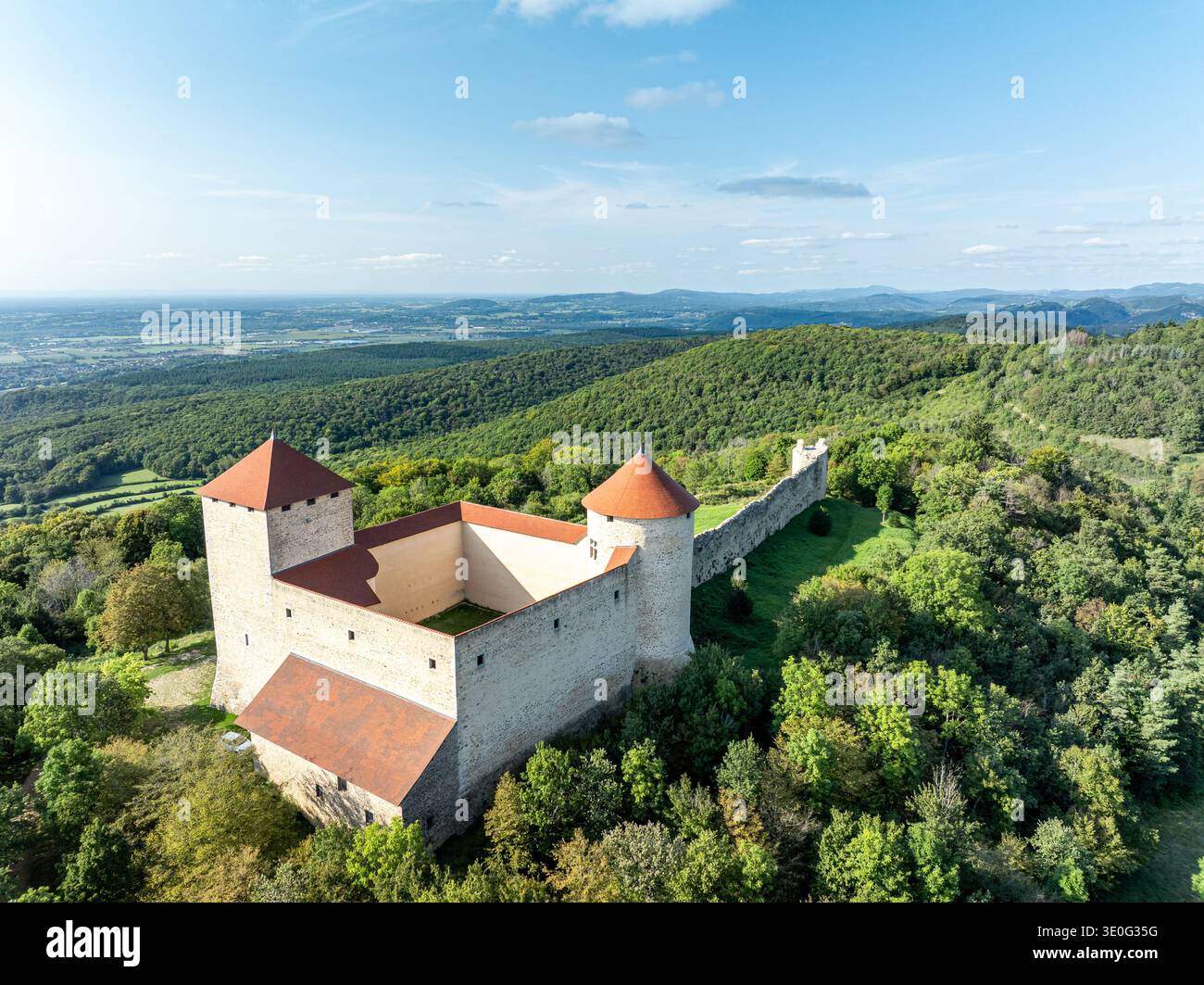 Vue aérienne d'une forteresse médiévale sur une colline avec des remparts en pierre blanche et des tours aux toits rouges, dominant une forêt verte dense, près d'Amberieu, Ain, France Banque D'Images