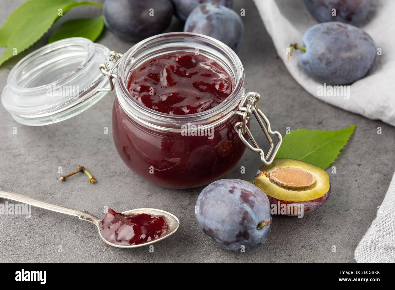 Confiture ou marmelade de prunes sucrées dans un pot en verre closeup et fruits frais crus entiers et tranchés avec des feuilles sur fond de table en pierre gris foncé. Somme saisonnière Banque D'Images