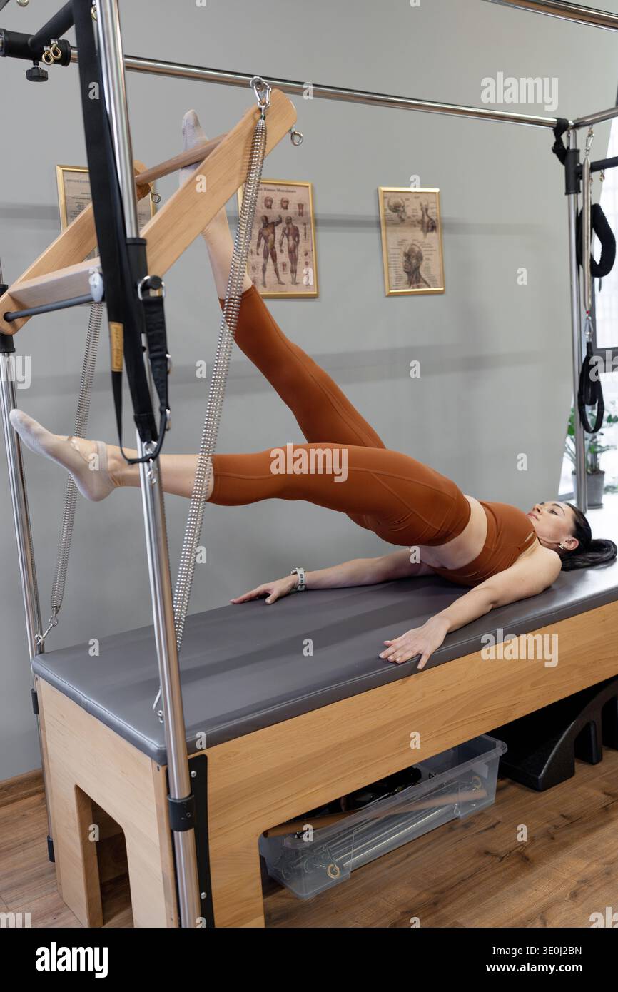 Femme faisant pilates se déplace avec de larges jambes et bras sur la table trapèze dans l'espace studio ensoleillé. Pilates bouge, jambes larges, table trapèze, studio ensoleillé, bras Banque D'Images