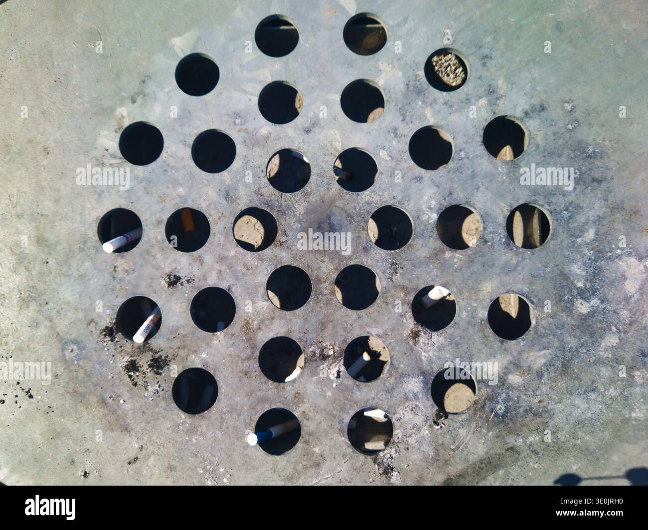 Vue panoramique d'un couvercle de cendrier circulaire en métal. La surface tachée et altérée présente un motif géométrique de trous avec de la ciga rejetée Banque D'Images