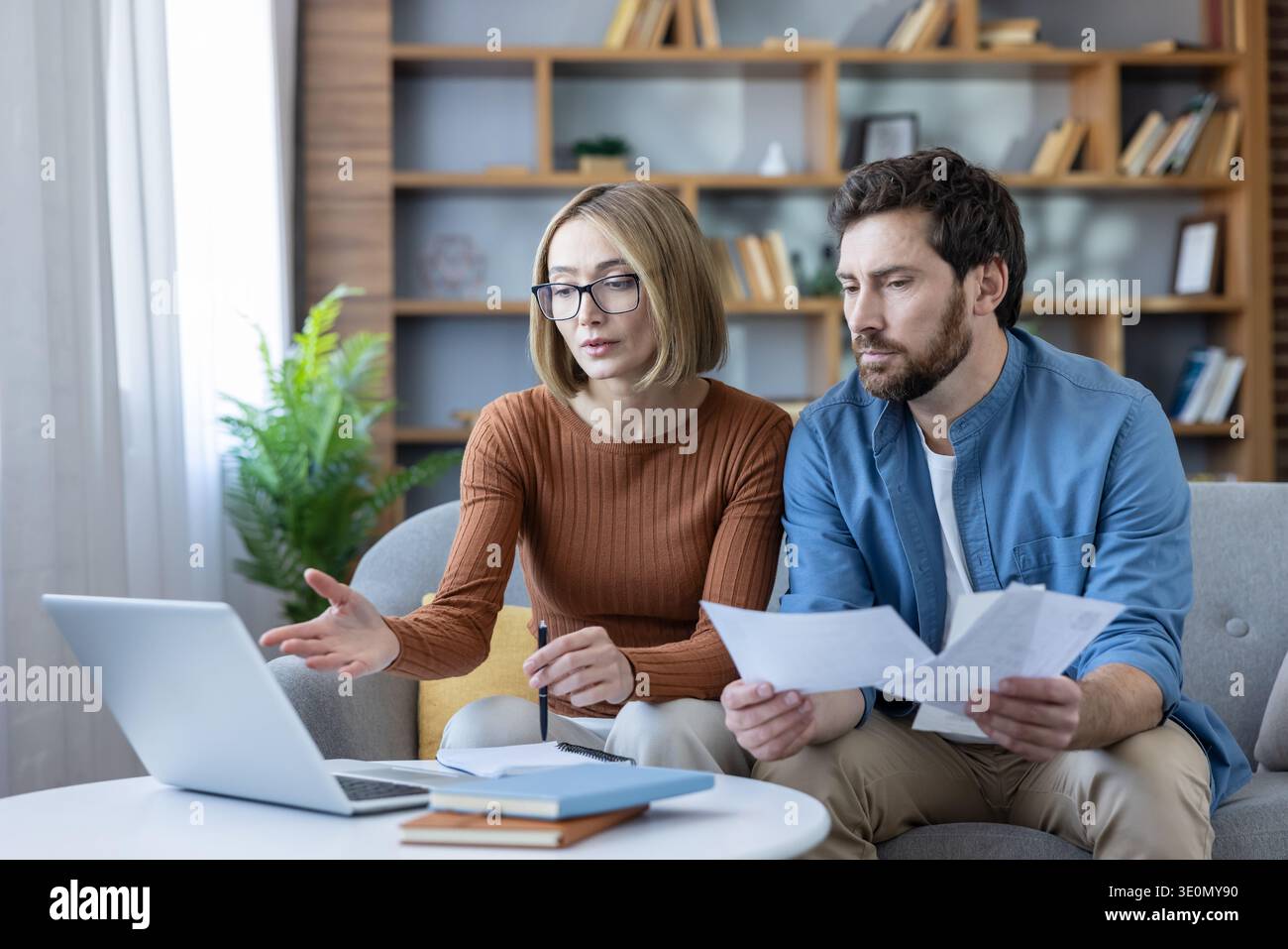 Couple analysant des documents financiers et utilisant un ordinateur portable pour la gestion de l'argent, la budgétisation et la planification des dépenses ensemble sur un canapé dans leur salon Banque D'Images