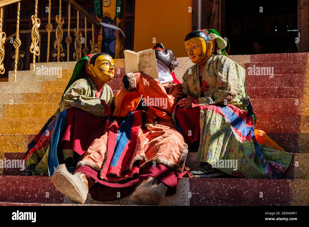 Des interprètes en masques traditionnels et robes florales étudient un texte religieux sur les marches pendant le festival Diskit Gustor. Banque D'Images