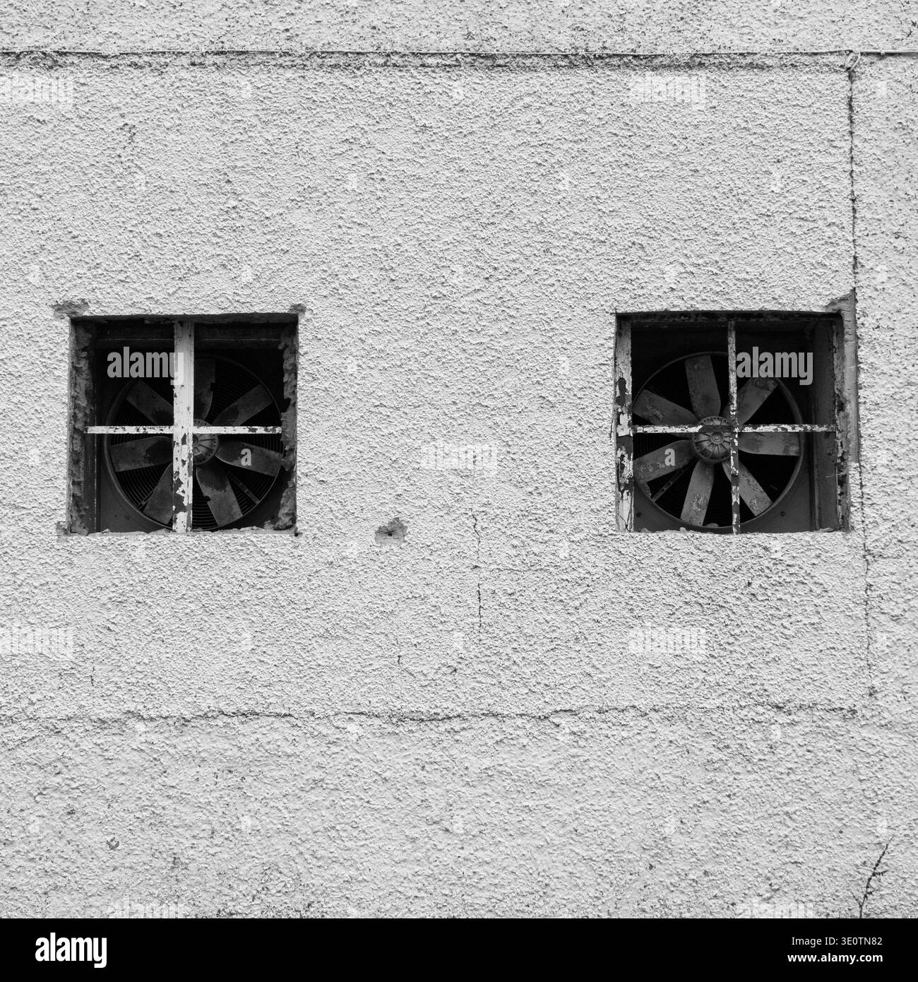 Deux ventilateurs industriels sont montés dans des ouvertures carrées dans un mur de béton rugueux et texturé. Les ventilateurs sont protégés par des grilles métalliques. L'image est en noir a Banque D'Images