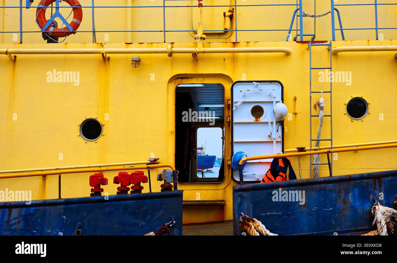 Vue latérale de la coque d'un navire jaune avec porte de cabine ouverte, bouée de sauvetage, échelles, hublots et équipement de sécurité sur balustrade bleue dans le port de Kaohsiung. Banque D'Images