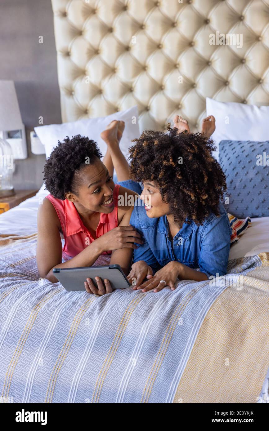 Deux femmes afro-américaines allongées sur le lit portant un haut corail et une chemise en denim, tenant une tablette Banque D'Images