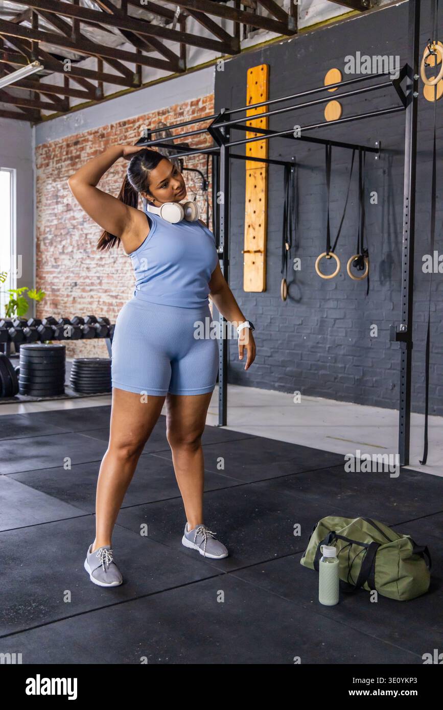 Femme afro-américaine adulte étirant le cou dans un studio de fitness portant une tenue bleue près de la plate-forme de pull-up Banque D'Images
