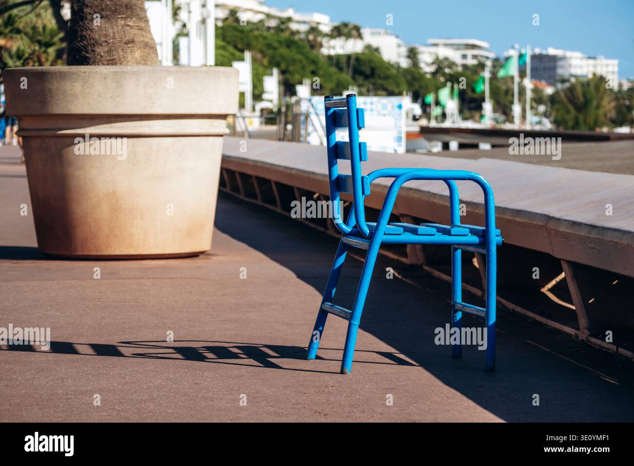 Chaise en métal bleu vif projetant une longue ombre sur le trottoir ensoleillé le long de la promenade de la Croisette à Cannes Banque D'Images