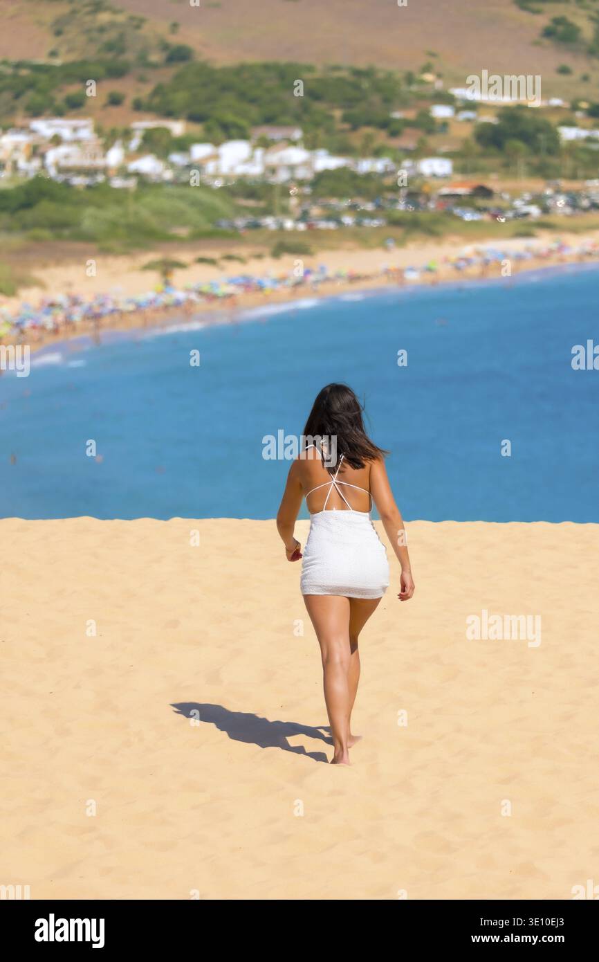Jeune femme marchant sur une dune de sable vers la plage de bolonia, avec des eaux turquoises et une plage pleine de parasols colorés, à tarifa, cadix, andalousie, s. Banque D'Images