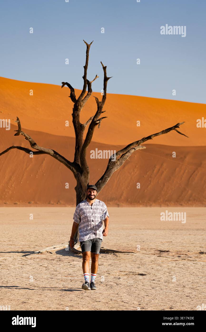 Un voyageur solitaire marche à travers le paysage sombre de Deadvlei, en Namibie, avec un arbre mort solitaire et des dunes de sable orange vibrantes sous un ciel bleu clair Banque D'Images