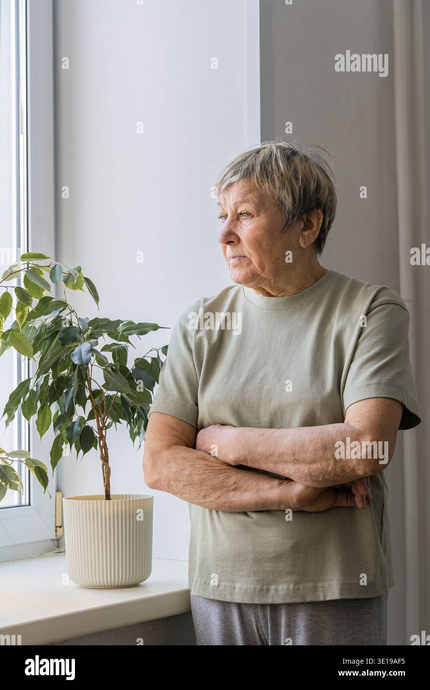 Femme senior debout par la fenêtre avec les bras croisés, réfléchie et pensive dans la lumière calme de la maison, reflétant sur la solitude et la mémoire Banque D'Images