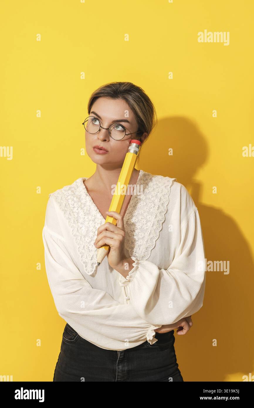 Pensive jeune femme portant des lunettes tenant grand crayon sur fond jaune Banque D'Images