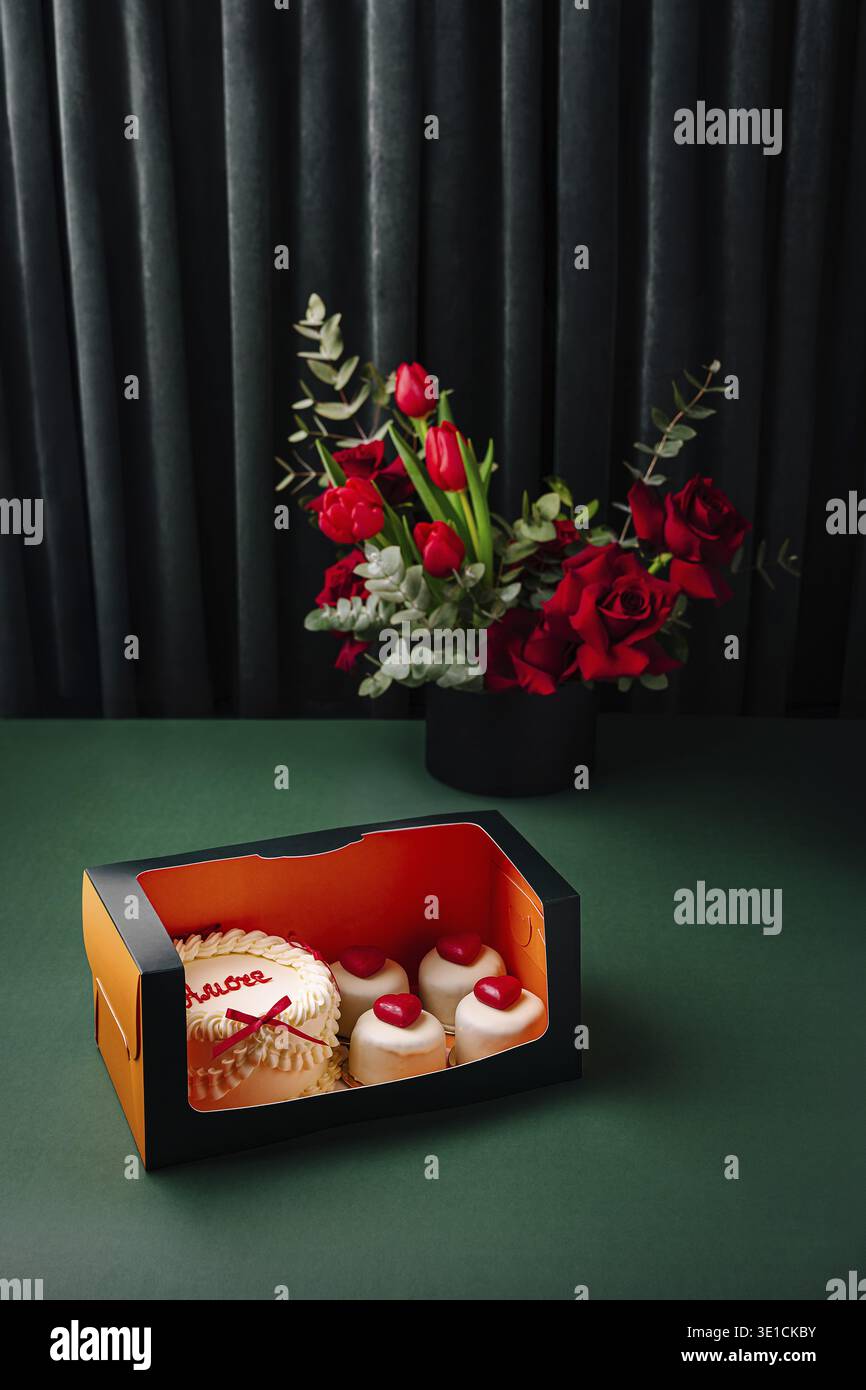 Une boîte magnifiquement agencée présente des cupcakes décorés avec des garnitures de coeur à côté d'un bouquet vibrant de fleurs rouges. La toile de fond est sombre, ele Banque D'Images