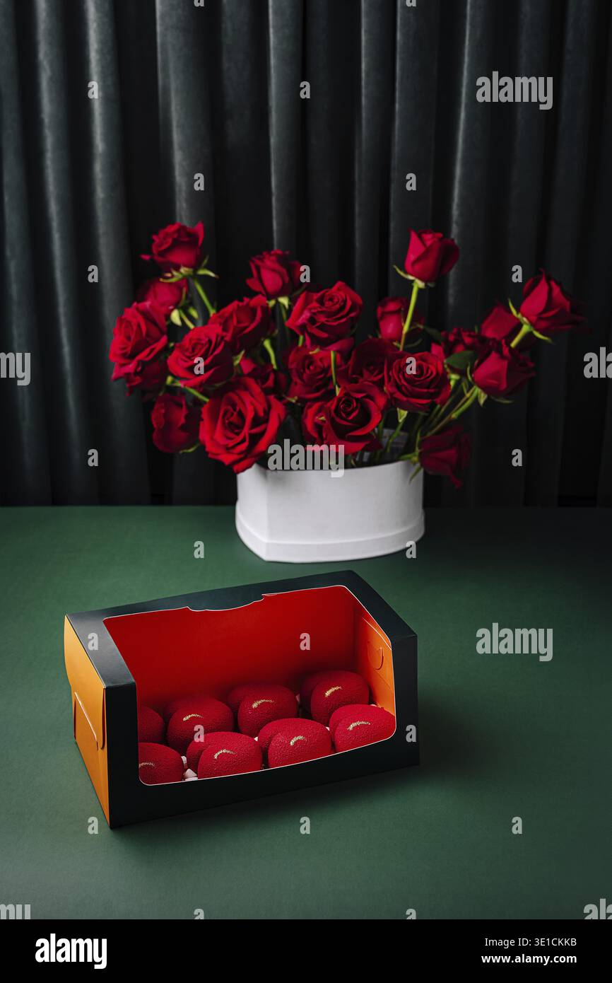 Un bel affichage présente des roses rouges vibrantes dans un pot blanc placé près d'une boîte colorée contenant de petits objets décoratifs. La toile de fond sombre améliore Banque D'Images