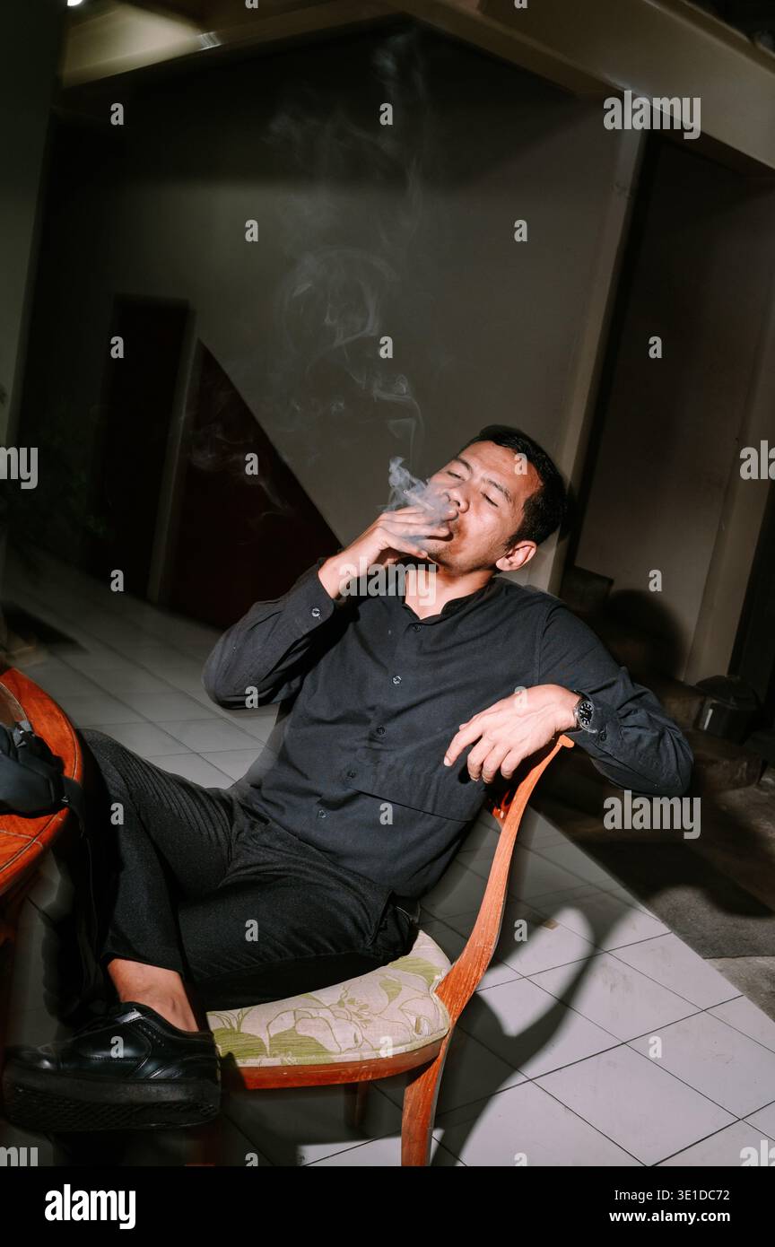 Un homme s'assoit confortablement, exhalant la fumée d'une cigarette à l'intérieur. L'éclairage dramatique met en valeur son moment de contemplation et de détente. Banque D'Images