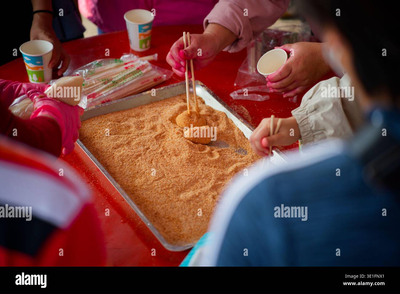 Gros plan des mains façonnant le mochi de riz collant dans un plateau de sable sur la table rouge traditionnelle, avec des tasses et des outils, capturant la préparation alimentaire Hakka à Taiwan. Banque D'Images
