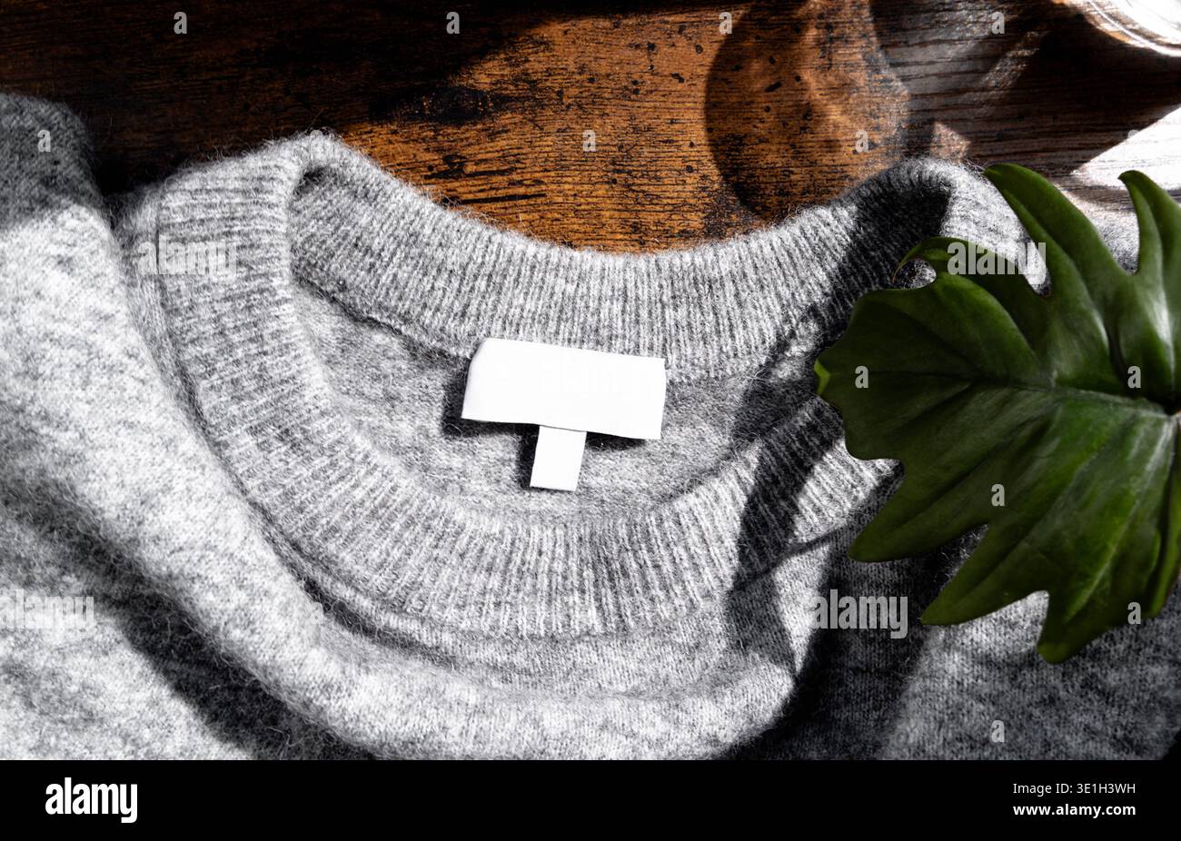 Pull tricoté gris avec étiquette de vêtement vierge sur une surface en bois et feuille verte. Concept de mode minimal avec texture naturelle, style confortable et marque Banque D'Images