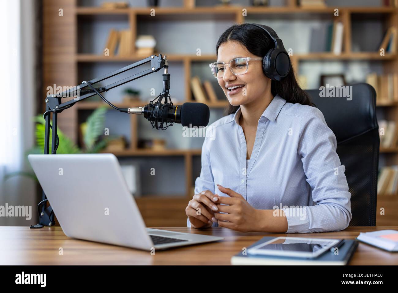 Jeune femme portant des écouteurs et des lunettes, parlant dans un microphone professionnel à un bureau avec un ordinateur portable, créant du contenu audio numérique pour sa diffusion en ligne Banque D'Images