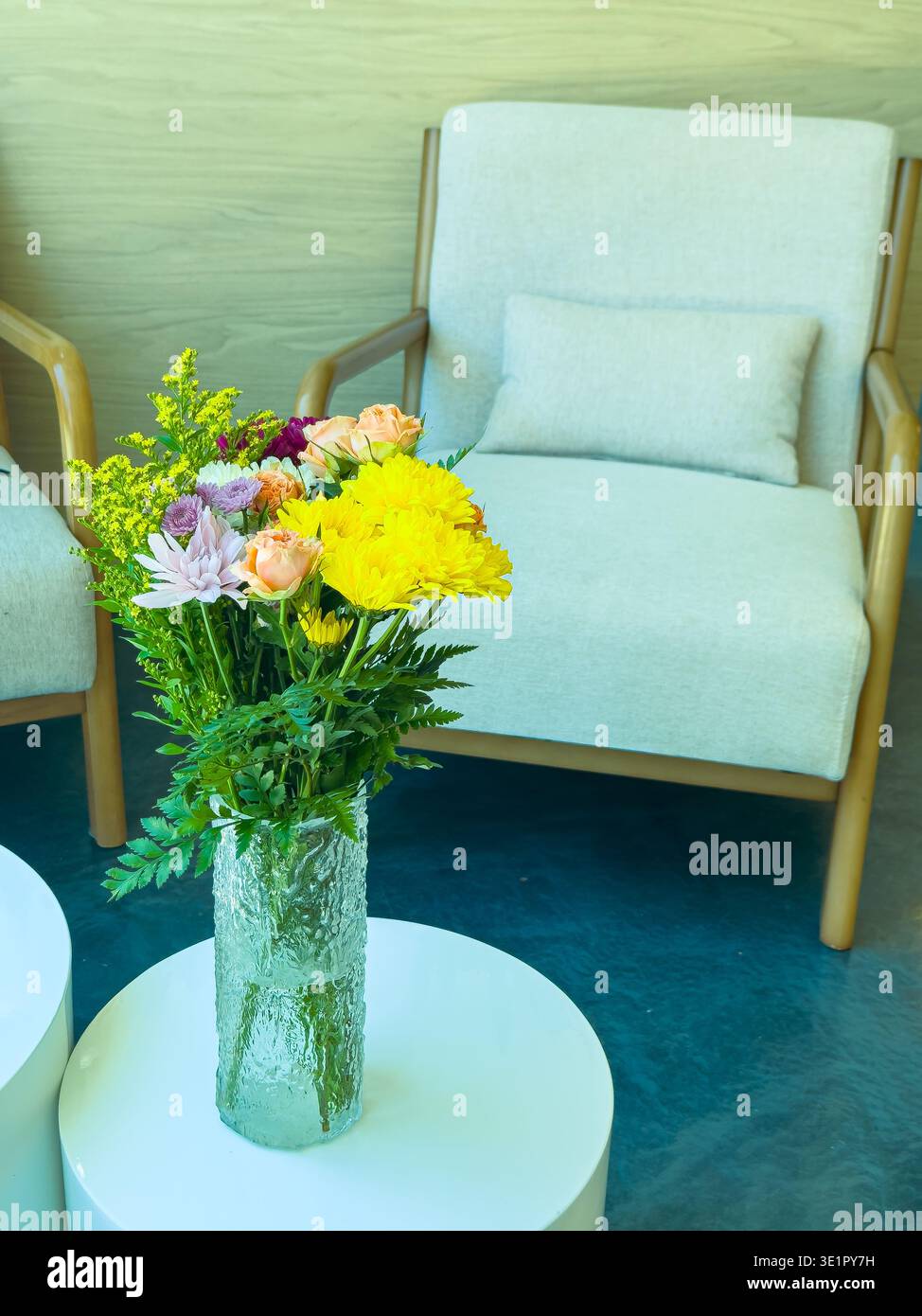 Un vase en verre contient une variété de fleurs, placées sur une table blanche à côté d'une chaise de couleur claire dans un espace intérieur bien éclairé. La lumière du soleil remplit la zone, hig Banque D'Images