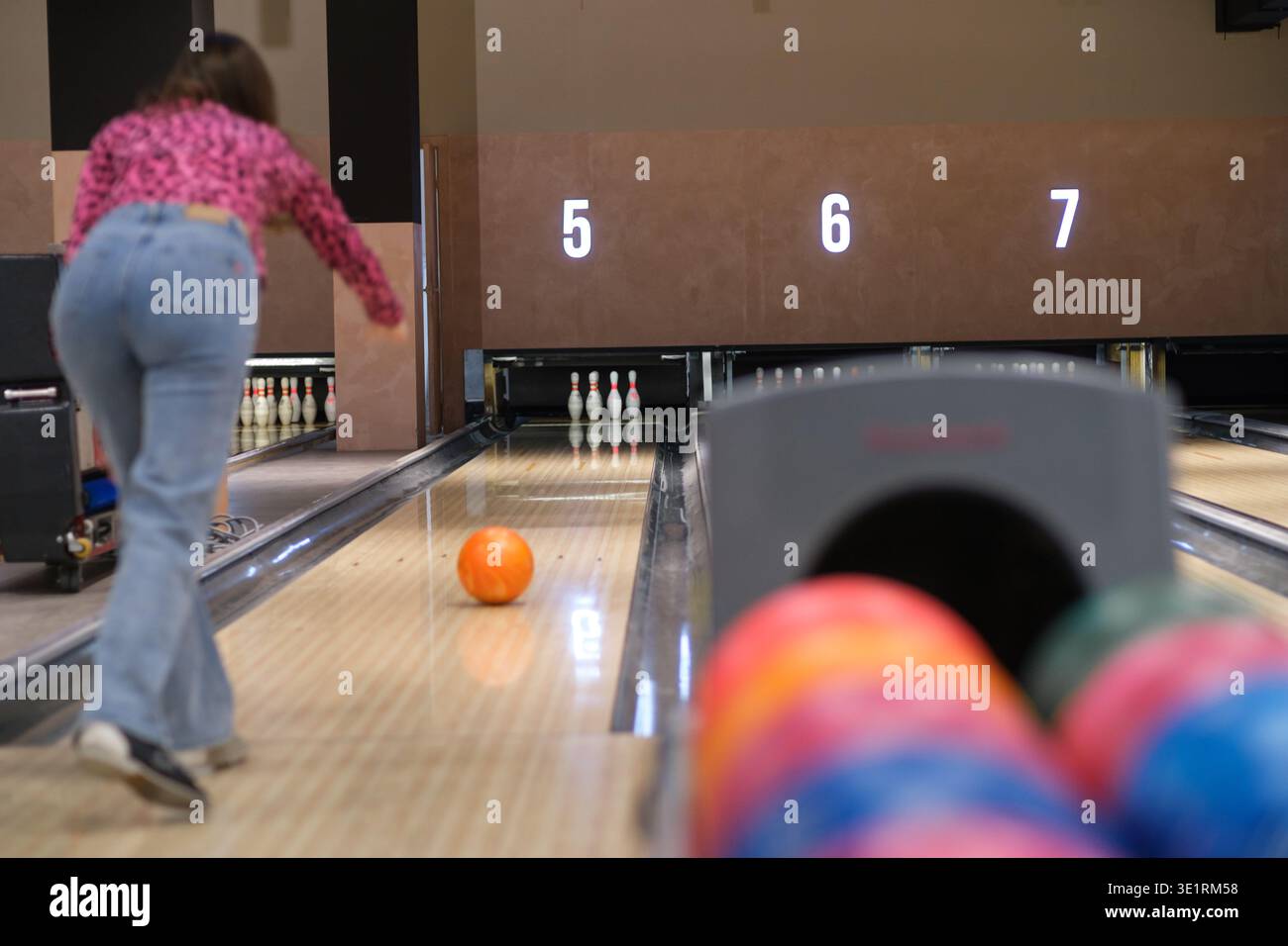 Jeune femme dans des bols de pull rose boule orange en bas de la piste de bowling, avec épingles et affichage de score visible dans le fond Banque D'Images