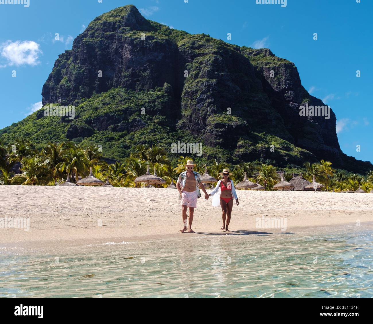 Un couple joyeux se promène main dans la main le long des rives d’une magnifique plage à Maurice, entourée d’une végétation luxuriante et de falaises majestueuses. Le Morne Maurice Banque D'Images