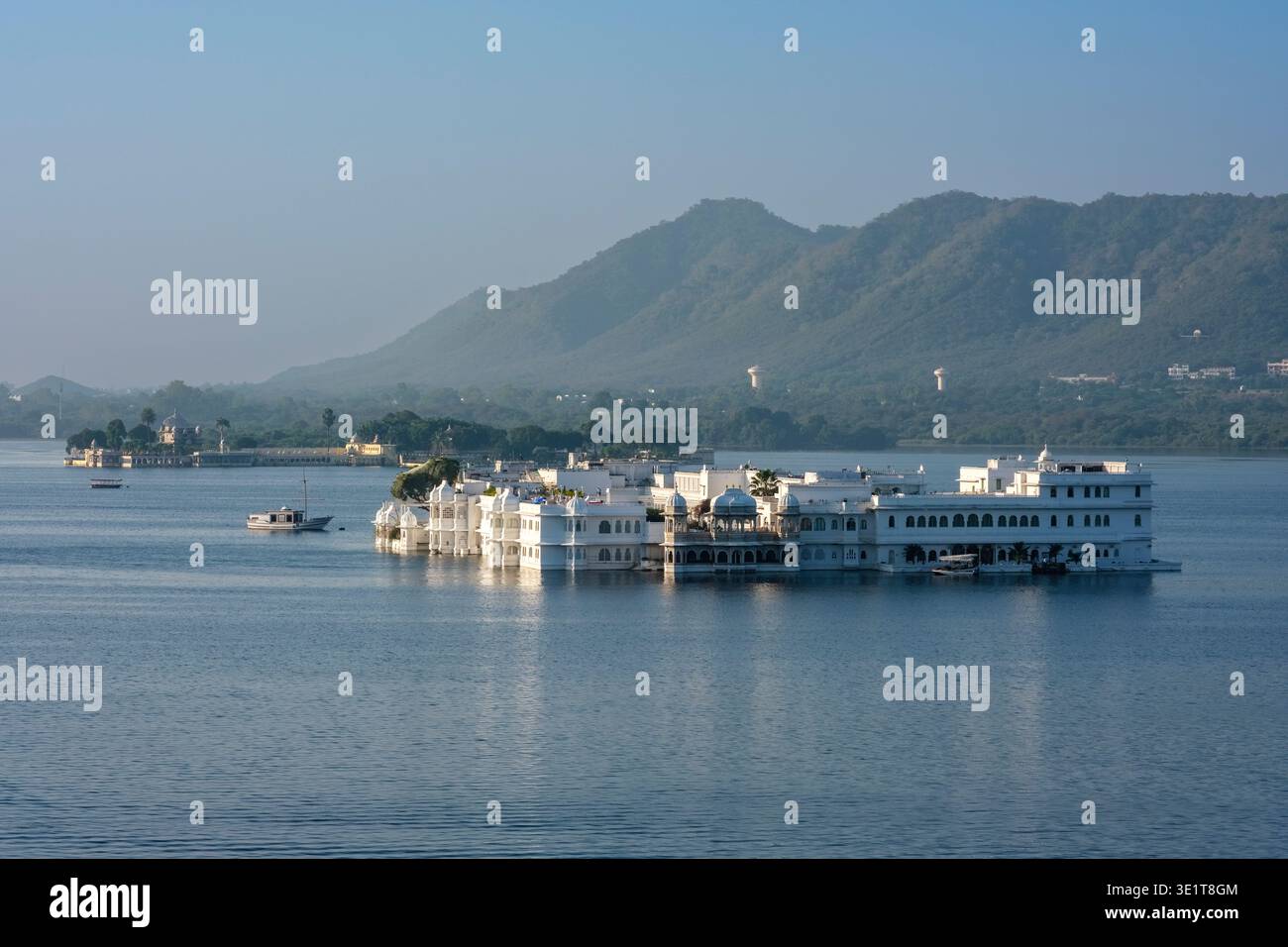 Le Taj Lake Palace en marbre blanc se trouve sur les eaux calmes du lac Pichola à Udaipur, avec les collines Aravali visibles à travers la distance brumeuse. Banque D'Images