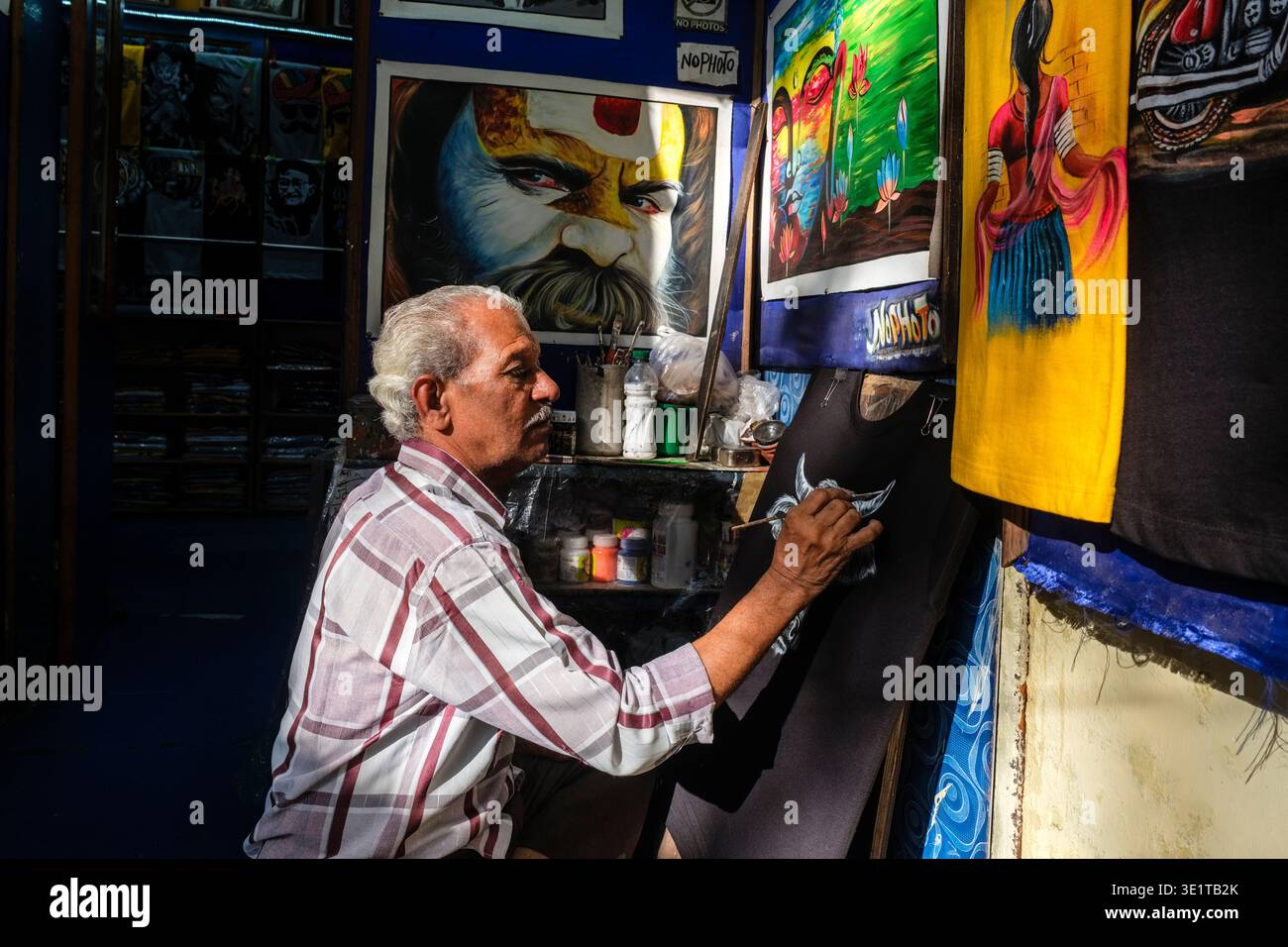 Dans un studio d'Udaipur, un artiste est assis les jambes croisées tout en peignant un taureau blanc sur un vêtement noir, entouré de diverses œuvres d'art colorées. Banque D'Images