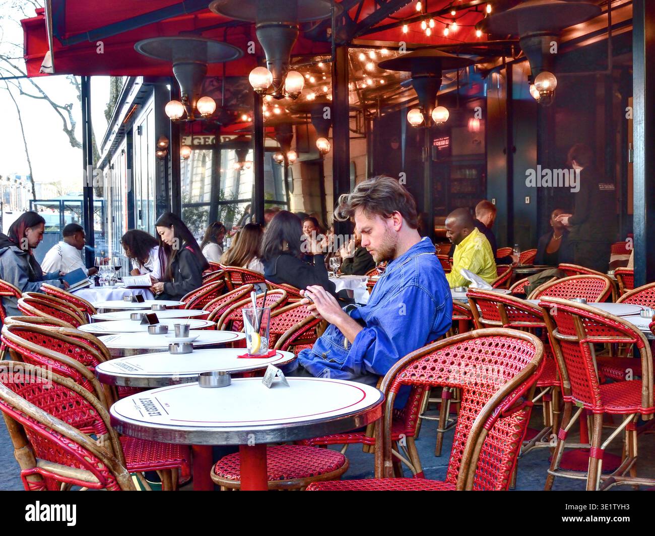 Jeune homme en chemise denim étudiant smartphone assis à la terrasse extérieure du George V Alma café - Paris 75008, France. Banque D'Images
