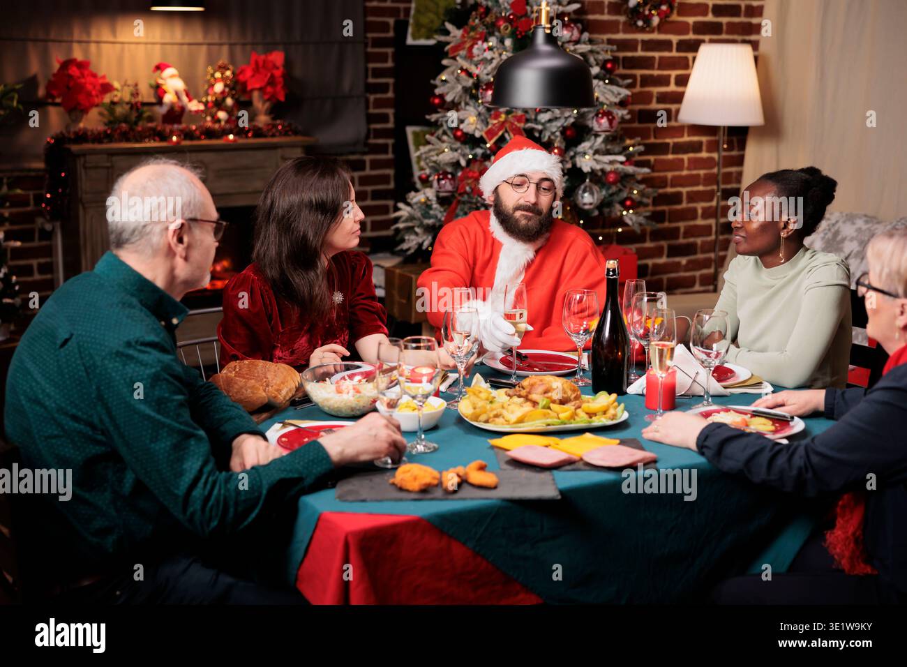 Homme en costume de père noël assis à table avec diverses personnes, profitant d'un dîner festif sur la fête de la veille de noël. Jeune en costume diffusant de la positivité pendant les vacances de décembre. Banque D'Images