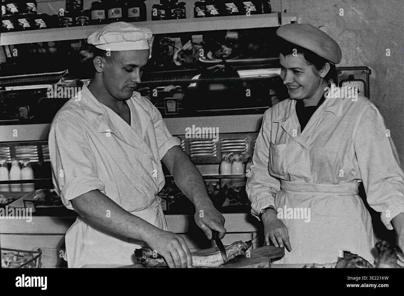 Authentique photographie d'archives en noir et blanc des années 1970 représentant un chef cuisinier masculin coupant habilement un grand morceau de viande ou de poisson rôti sur une planche de bois dans une cafétéria slavyansk. Une collaboratrice joyeuse dans un uniforme blanc et béret observe ses actions précises avec un sourire chaleureux. Ils se tiennent derrière un comptoir culinaire chargé de bocaux en verre traditionnels et de divers plats préparés. Cet instantané franc préserve la véritable mémoire du travail collectif et l'atmosphère d'un Donbass pacifique avant la guerre. Banque D'Images