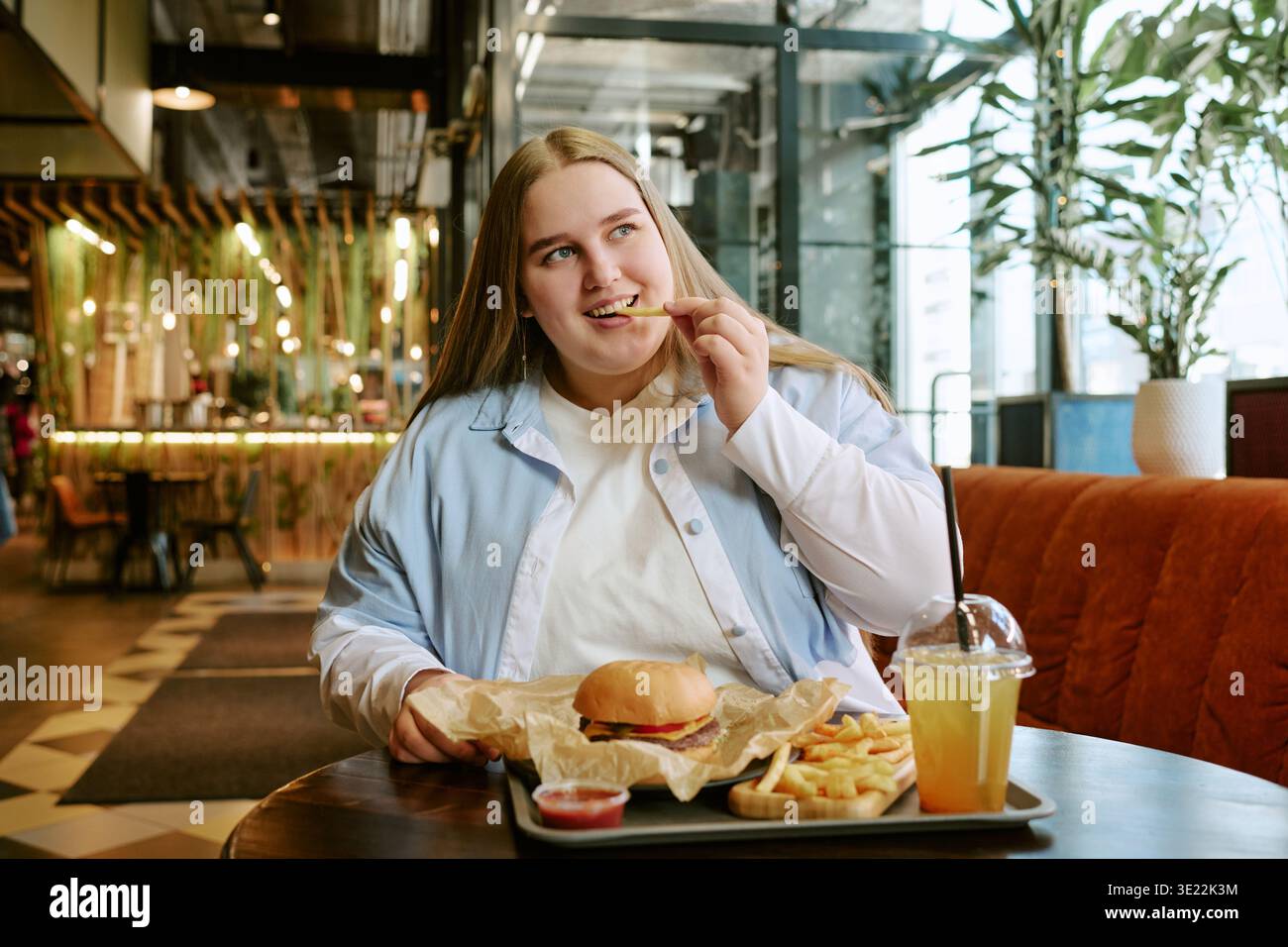 Jeune femme adulte mangeant des frites et un hamburger dans le café moderne Banque D'Images