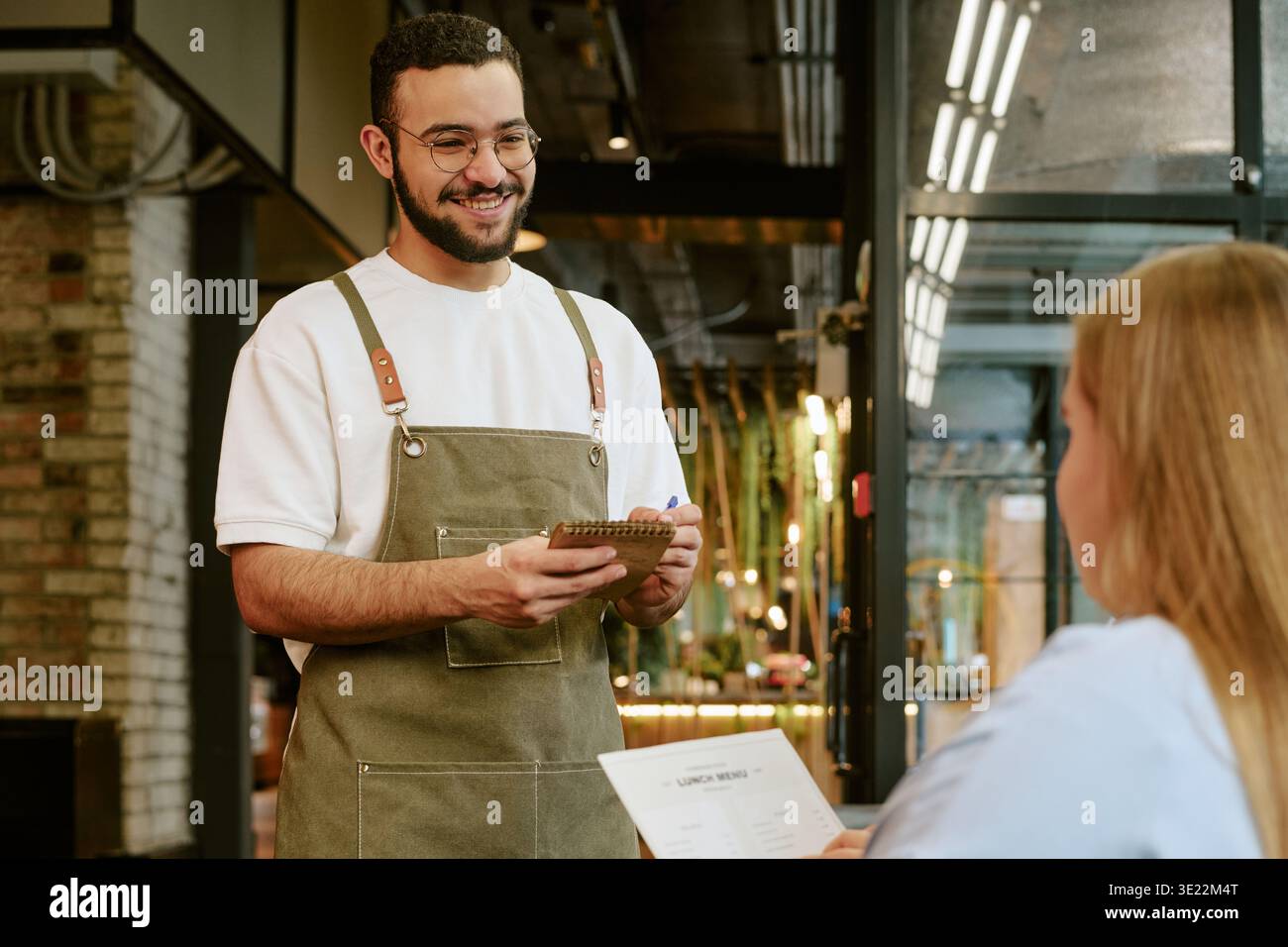 Jeune homme adulte souriant tout en prenant la commande d'une cliente Banque D'Images