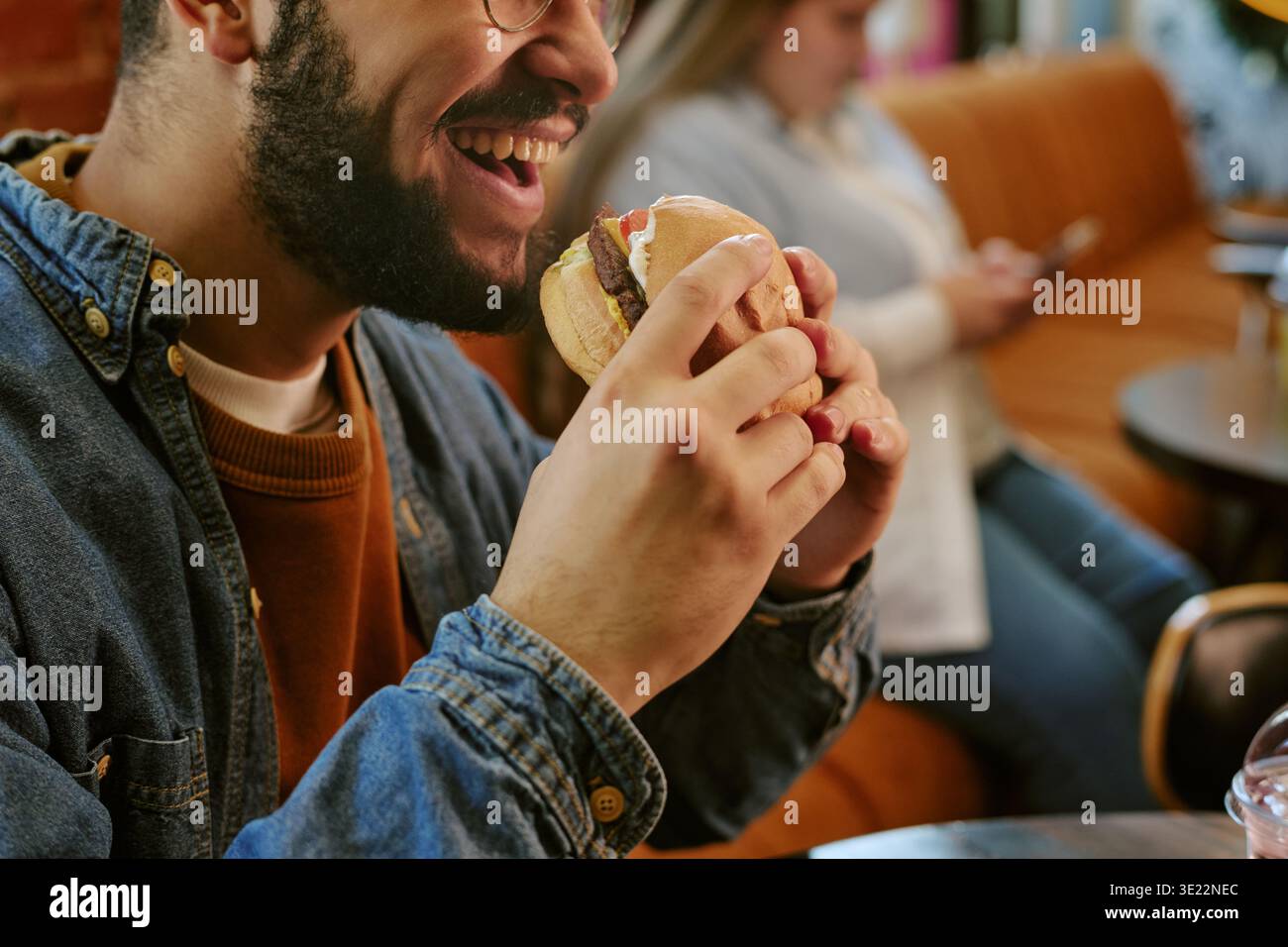 Jeune homme adulte souriant en mangeant un hamburger dans un cadre café Banque D'Images
