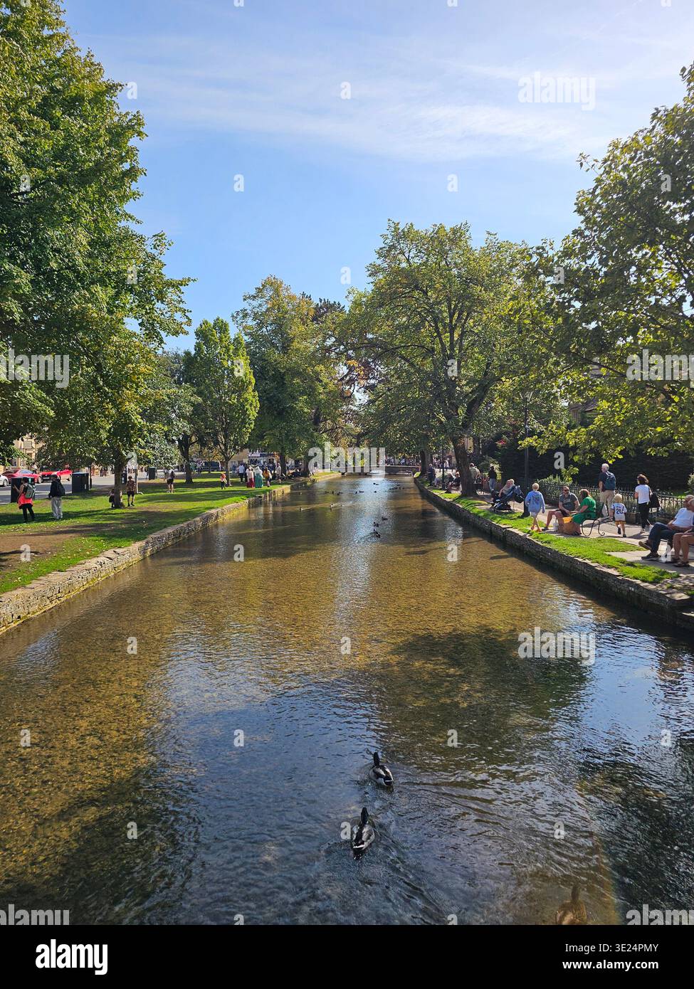 Une journée d'été bien remplie à Bourton-on-the-Water montre les visiteurs se détendre au bord du ruisseau, capturés depuis un pont au cœur des Cotswolds. Banque D'Images
