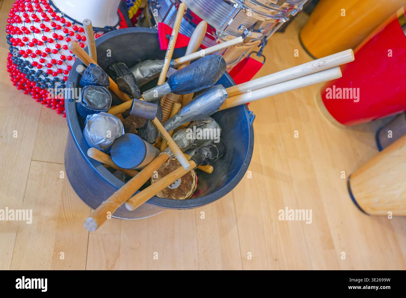 Un seau contenant différents types de maillets à percussion et de pilons placés sur un plancher en bois avec des instruments de musique colorés à proximité. Banque D'Images